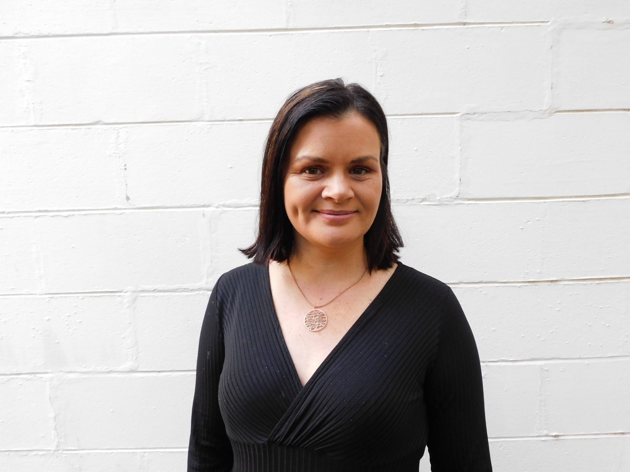 Portrait of Indigenous woman and activist Bianca Hunt. She stands in front of a white brick wall, smiling. She's wearing a black dress and pendant necklace.