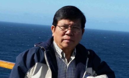 Mr Chau wears a jacket and glasses, and stands in front of the ocean.