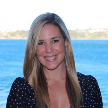 A portrait of Danielle Begg, founder of the Yoga Impact Charity. She's smiling wearing a black dress with white dots. The ocean is behind her.