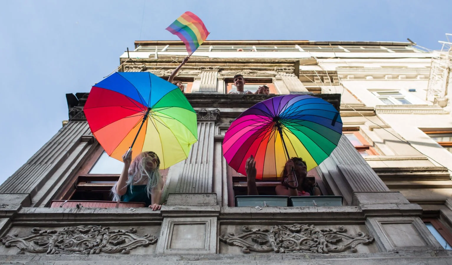 Umbrellas with the pride colours, held out of the window of a tall building. Blue sky is above.