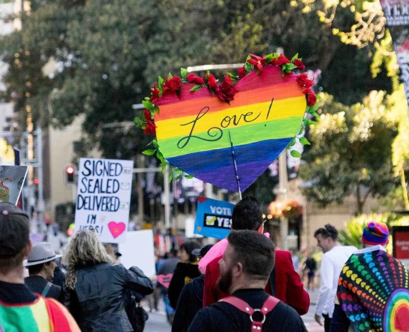 A photo of a crowd of people gather for a rally marriage equality rally in Sydney. There are numerous people holding rainbow flags and carrying signs that say 'Love!' and 'Vote Yes!'
